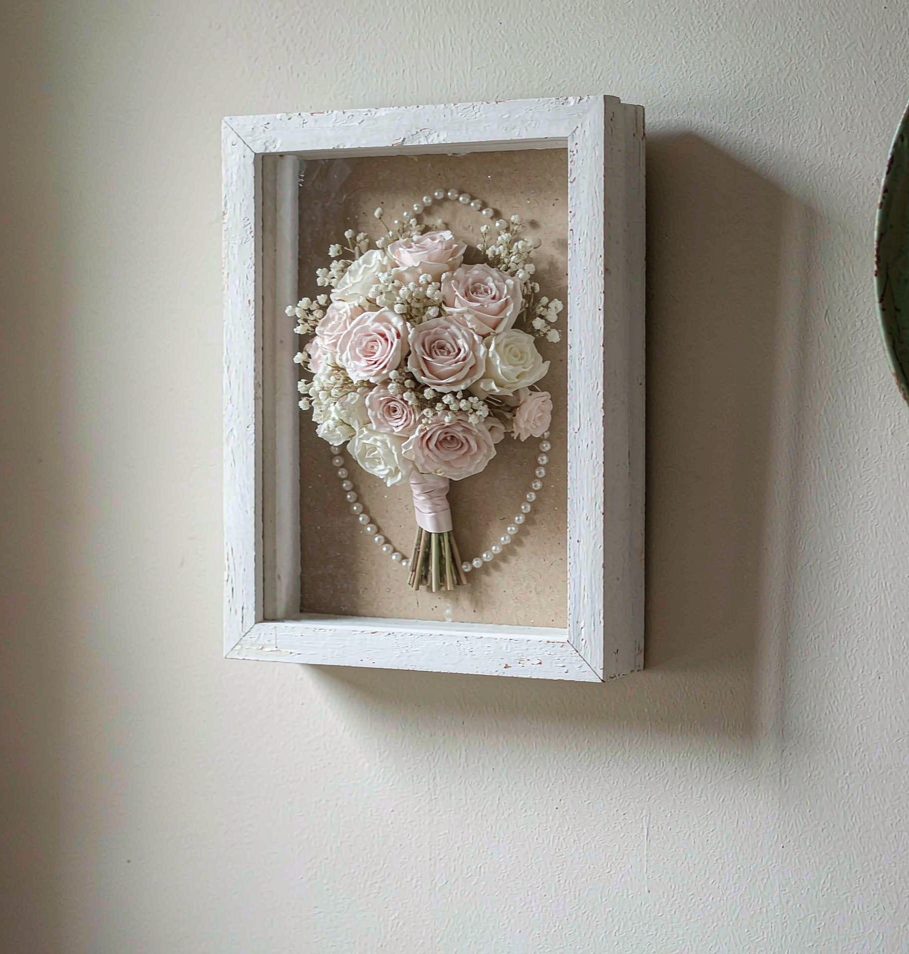 A preserved wedding bouquet in a white wooden shadow box frame hanging on an ivory wall.
